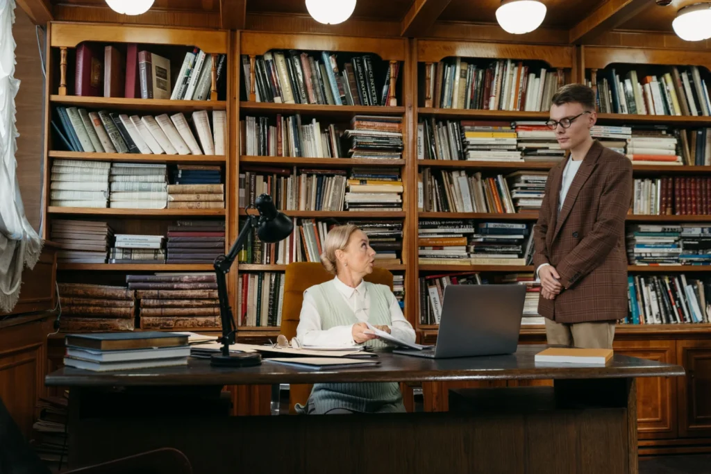 Dos personas conversando en una biblioteca universitaria junto a los estantes